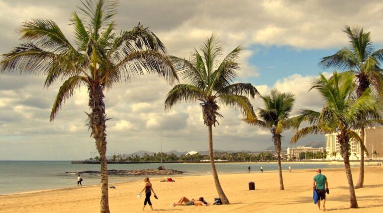 El turismo en Lanzarote y la búsqueda de alojamientos tranquilos en entornos volcánicos - - 779951 - Finca Tamaragua | Paradores.tv El turismo en Lanzarote y la búsqueda de alojamientos tranquilos en entornos volcánicos - Sunlit sandy beach with tall palm trees, people relaxing on the shore, and a calm sea under a partly cloudy sky.
